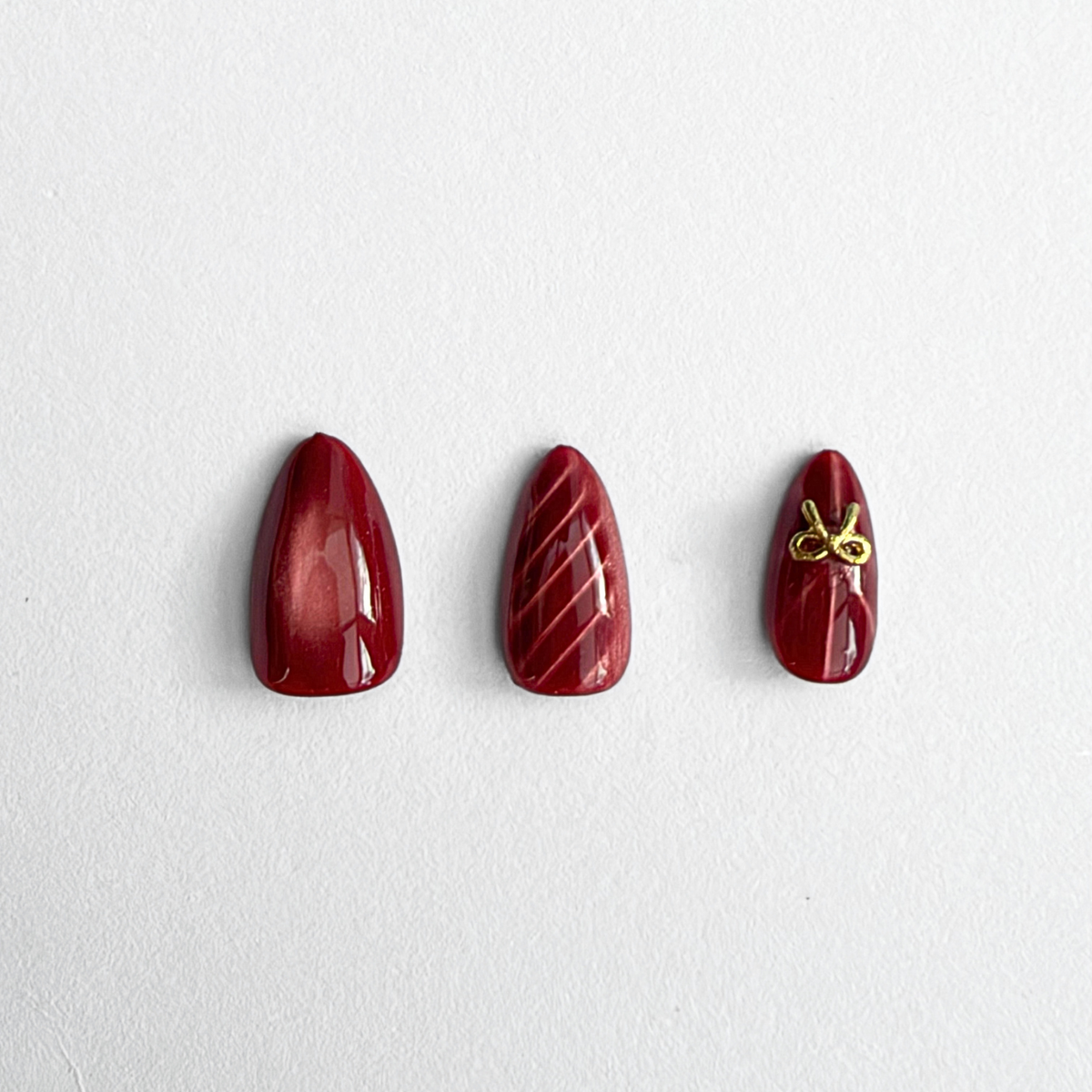 Deep red soft gel press-on nails, glossy finish, diagonal ribbed texture, gold bow accent nail