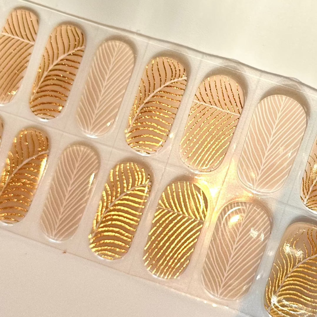Nude gel nail wraps with gold feather pattern and geometric designs for elegant manicure