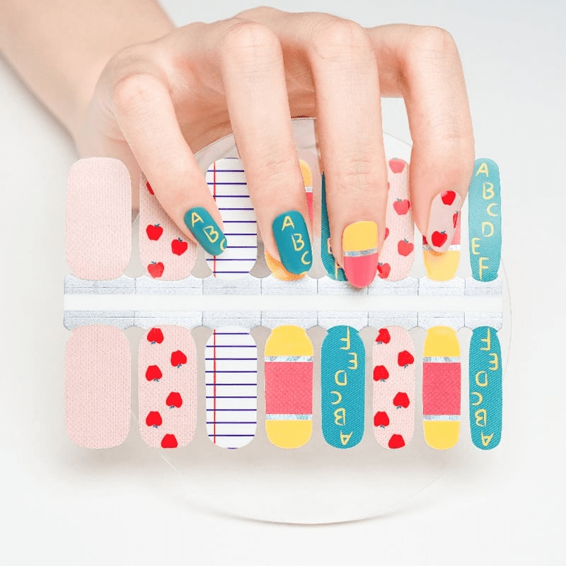 Hand wearing school-themed nail wraps with red apples, stripes, and teacher text patterns