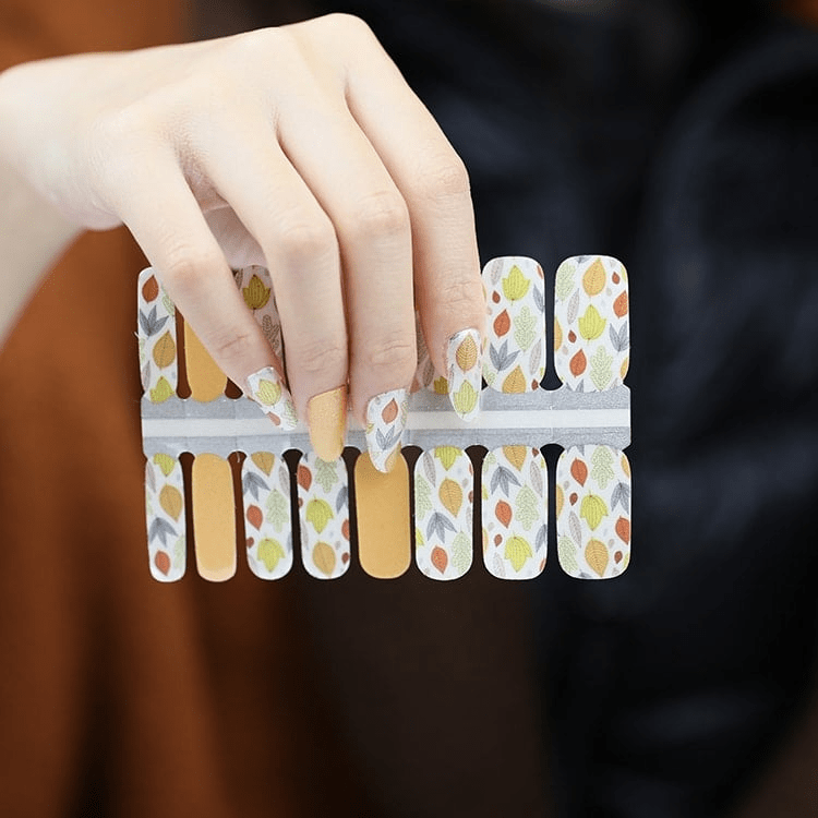 Hand holding autumn leaf pattern nail wraps with orange and yellow botanical designs