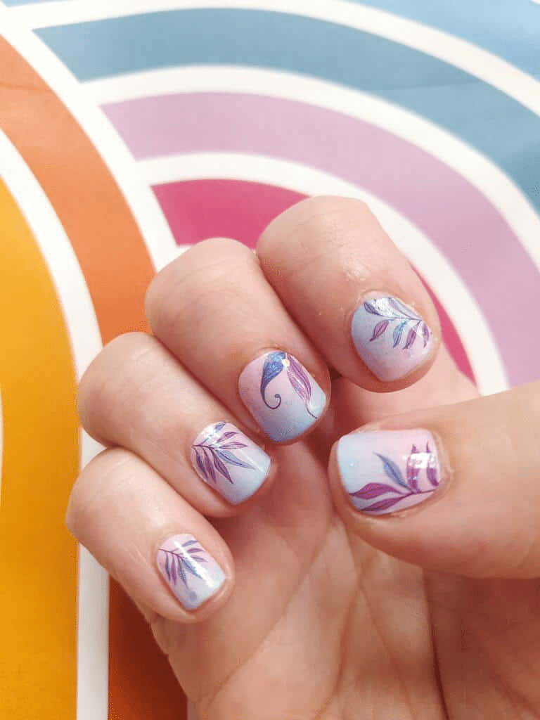 Hand wearing pastel gradient nail art wraps with purple botanical leaf designs over rainbow backdrop