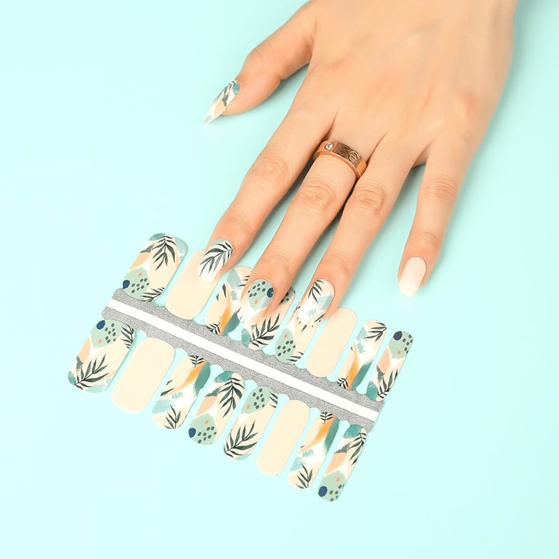 Hand with nail wraps sheet showing botanical leaf patterns in beige and dusty blue colours