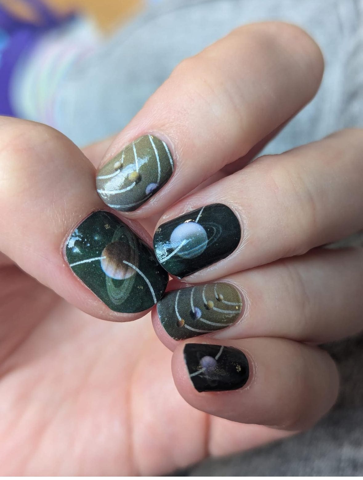 Space-themed nail wraps on hand featuring planets and metallic silver accents in dark colours