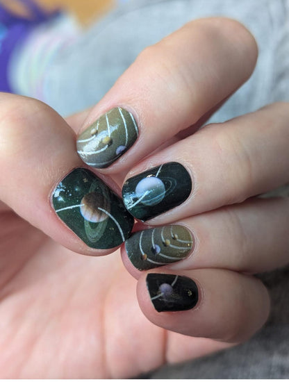 Space-themed nail wraps on hand featuring planets and metallic silver accents in dark colours