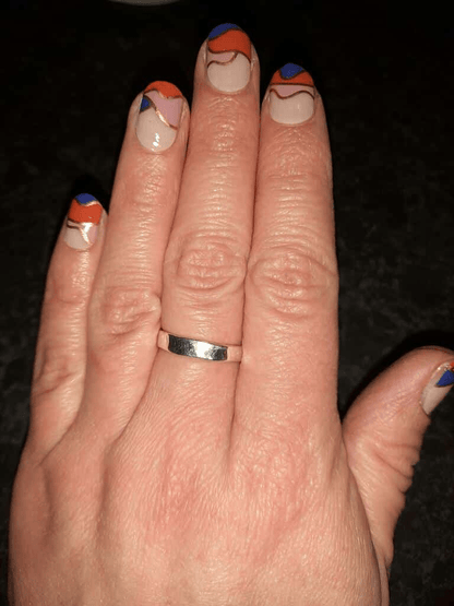 Hand with wedding ring displaying geometric nail wraps in navy blue, orange and nude design