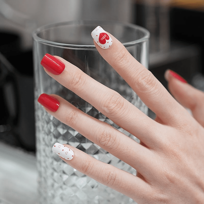 Red and white heart nail wraps with XOXO pattern displayed on hand near glass