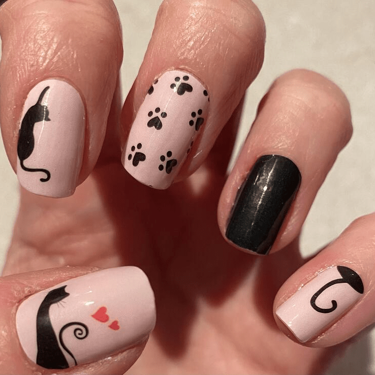 Pale pink nail wraps with black cat silhouettes, paw prints, hearts and a solid black accent nail
