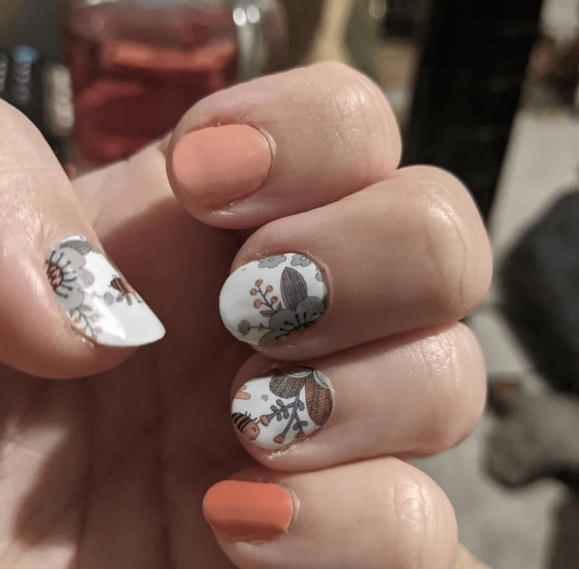 Close-up of peach and white floral bee nail wraps on hand with vintage botanical pattern