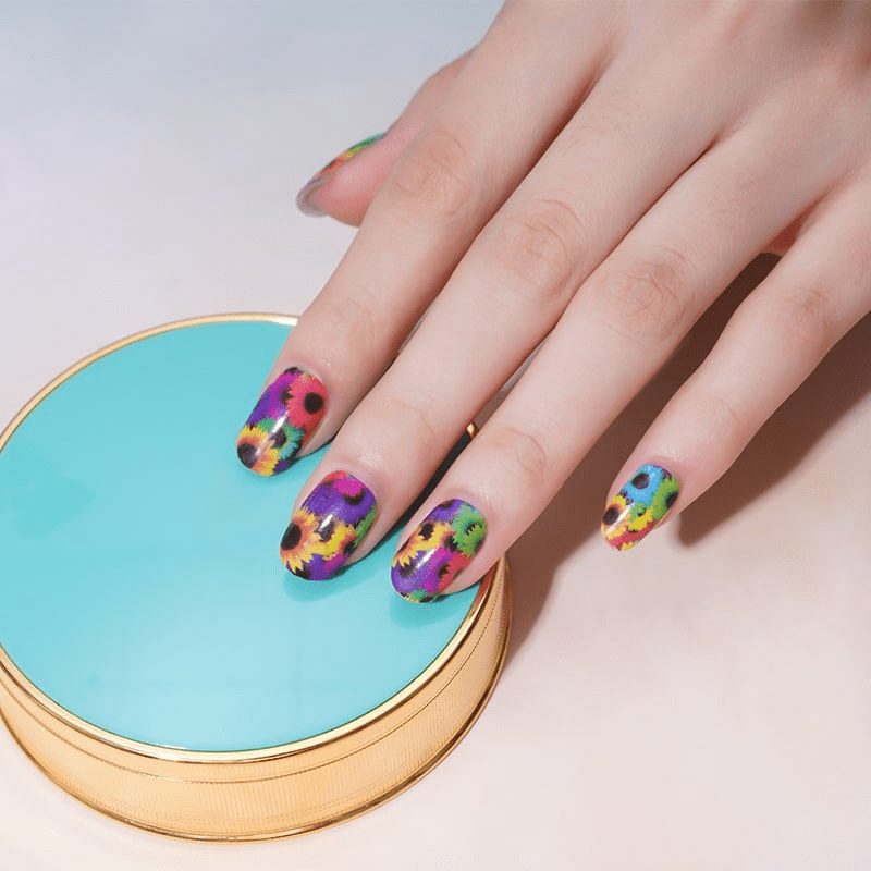 Hand displaying vibrant multicoloured sunflower nail wraps with rainbow floral patterns on blue surface