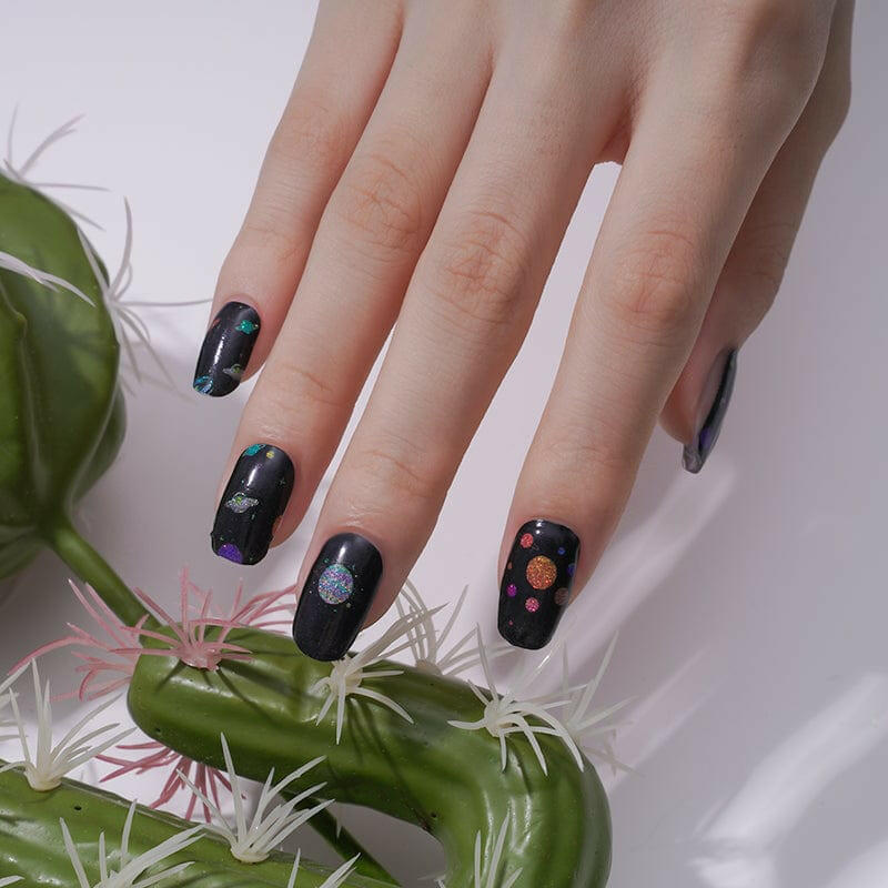 Space-themed nail art wraps with galaxy planets pattern on natural nails beside plant