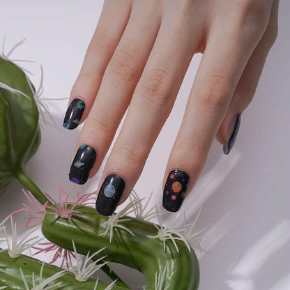 Space-themed nail art wraps with galaxy planets pattern on natural nails beside plant