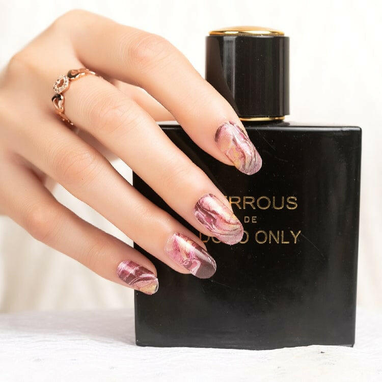 Hand wearing pink marble nail wraps with metallic accents holding black perfume bottle