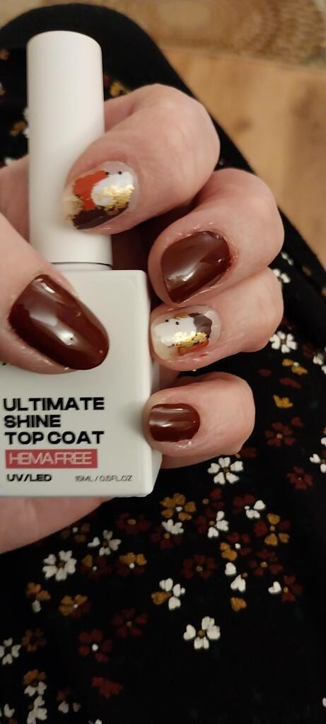 Hand wearing glossy burgundy nails with gold leaf accent and hema-free top coat bottle