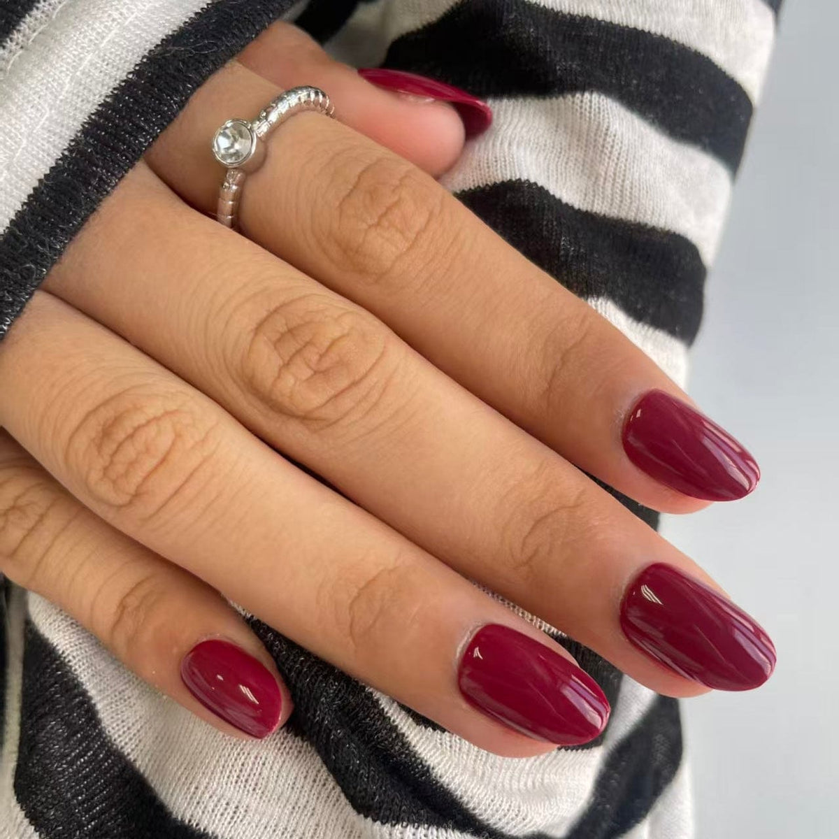 Deep burgundy red glossy short oval press-on nails on hand with striped jumper