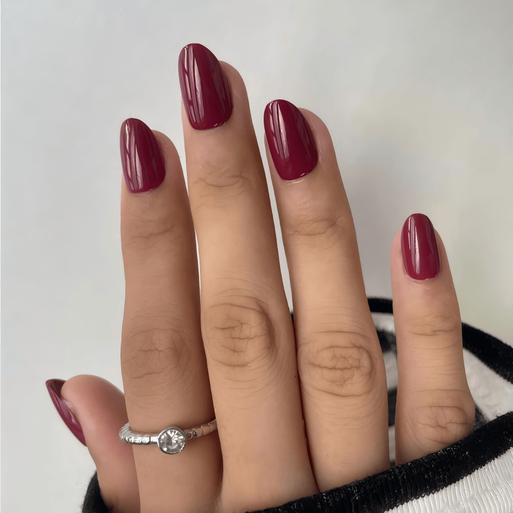 Deep burgundy glossy press-on nails on hand showing short oval shape gel nail extensions