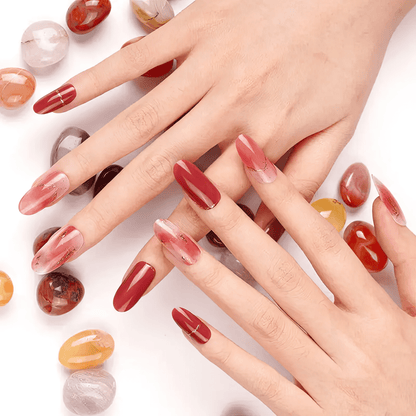 Hands displaying red burgundy chrome gel nail wraps with gold chain detailing surrounded by gemstones