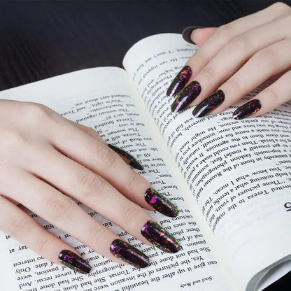 Hands with dark sparkly gel nail wraps holding open book showing gothic nail art